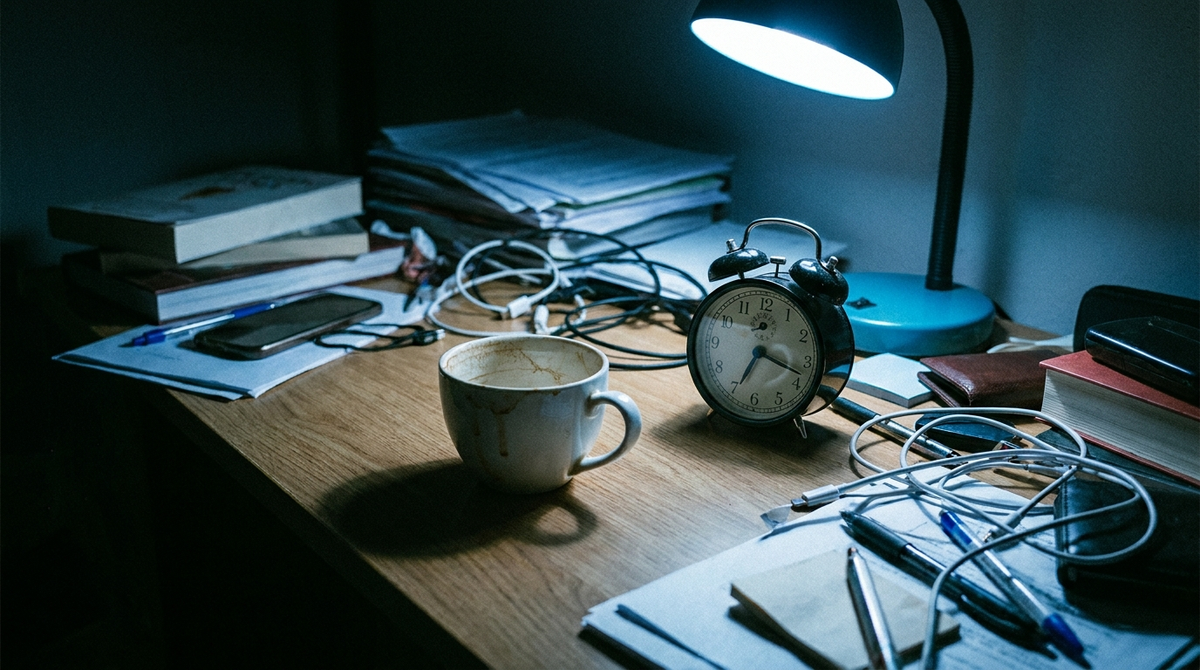 An empty coffee cup and alarm clock on a cluttered desk at night